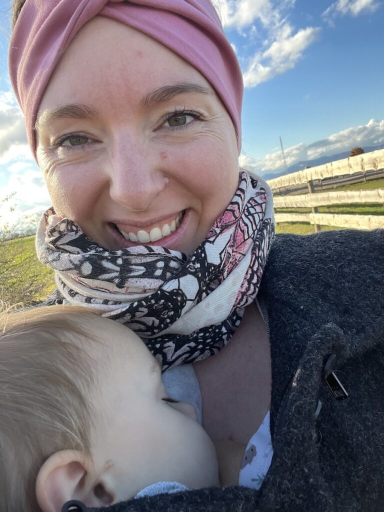 Lächelnde Frau mit Baby im Arm draußen auf einer Wiese unter blauem Himmel.